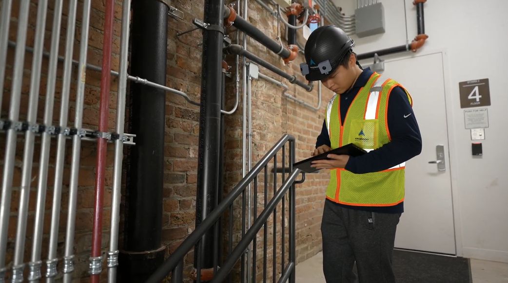 A worker wearing the Brilliant Builders helmet reviews results on a tablet in a stairwell